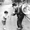 Vintage black‑and‑white photograph of a one‑man‑band street performer interacting with a young child on a quiet tree‑lined street.